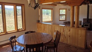 Un intérieur rustique avec une table à manger en bois, des chaises, un coin bar et de grandes fenêtres donnant sur l’extérieur.