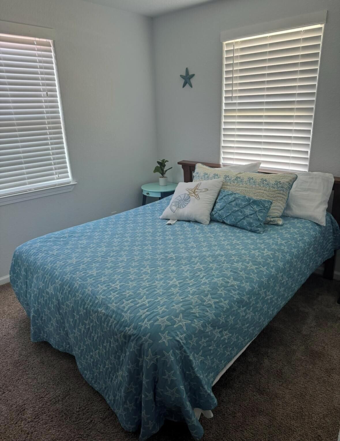 A bedroom with a bed, a nightstand, a wall star decoration, and two windows with blinds.