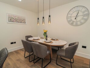 Un coin repas comprenant une table ronde, quatre chaises, une grande horloge murale, des luminaires suspendus et un tableau représentant des fleurs.