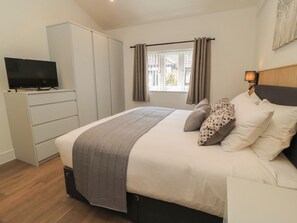 Une chambre à coucher avec un lit, un téléviseur posé sur un meuble, une fenêtre avec des rideaux et une tête de lit agrémentée d’une lampe.