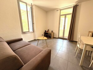 A living room with a sofa, a small round table, a dining table with chairs, and a sliding glass door. - Apartment T2 Corte center (Corte)
