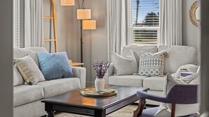 Ein Wohnzimmer mit einer Couch, einem Couchtisch und einem Beistelltisch. Ein Fenster mit Jalousien, eine Lampe und eine Vase mit Blumen auf dem Couchtisch.