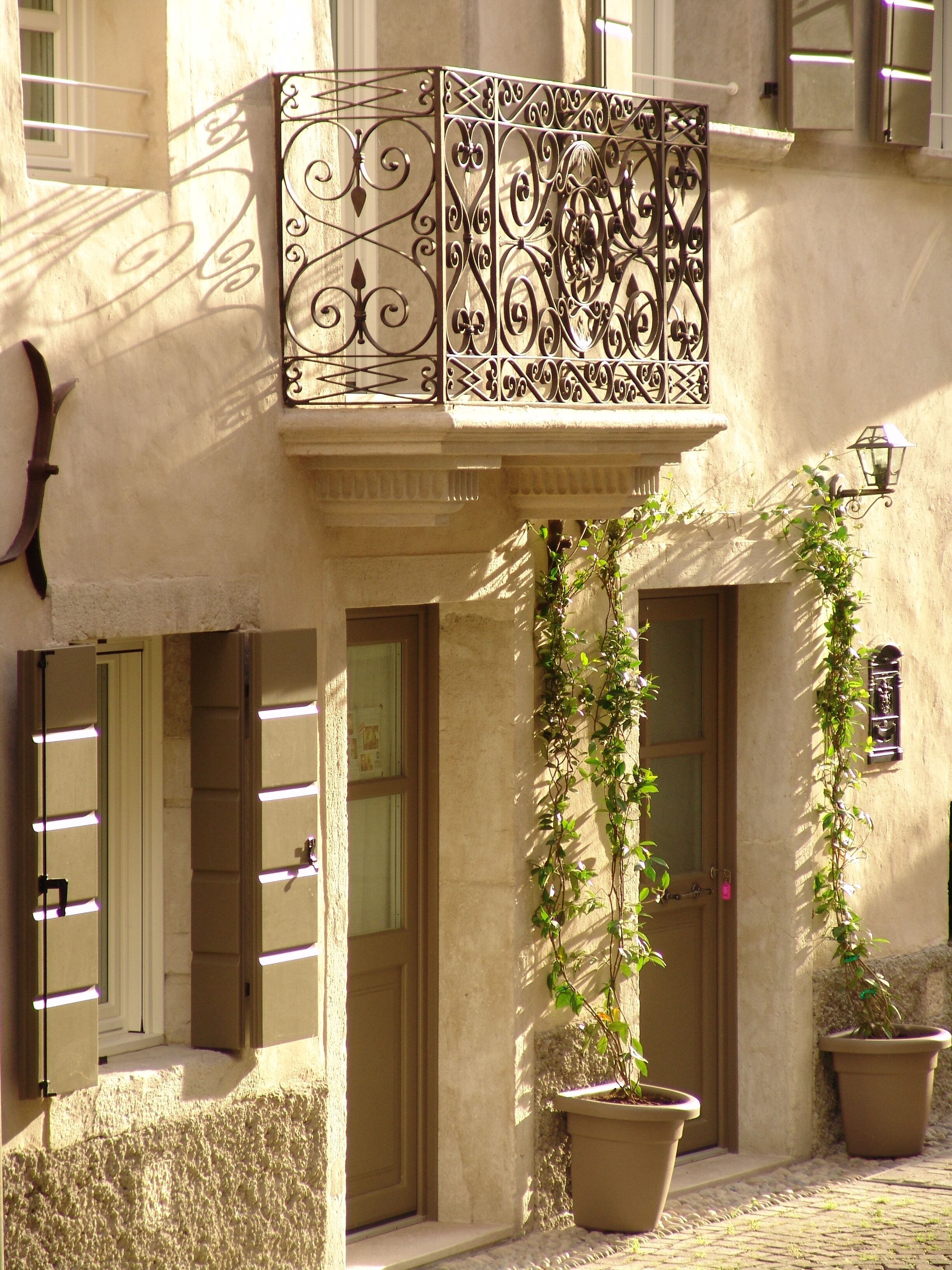 A building with a balcony featuring ornate metalwork, potted plants, and a door with a window.