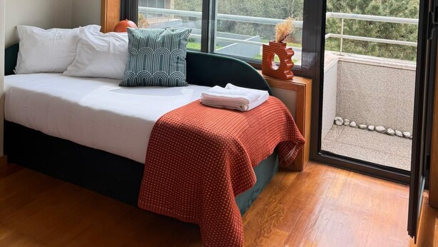 Une chambre à coucher avec un lit, des oreillers, une couverture rouge, une table de chevet et une vue sur les arbres à travers de grandes fenêtres.