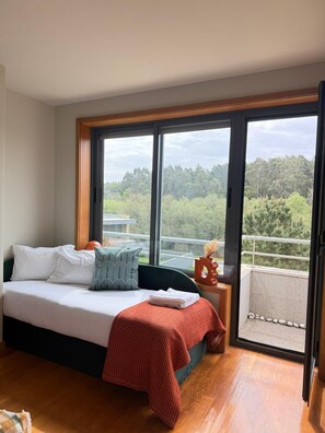 A bedroom with a bed, pillows, a red blanket, a nightstand, and a view of trees through large windows. - Pedra Moura Terrace by Porto Stories (Vila Nova de Gaia)