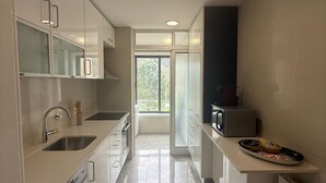 Une cuisine moderne dotée d’un évier en acier inoxydable, de meubles blancs, d’un four à micro-ondes et offrant une vue sur un balcon grâce à une porte vitrée.