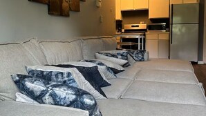 Una sala de estar moderna con un sofá seccional, una zona de cocina con electrodom ésticos de acero inoxidable y una obra de arte colgada en la pared.