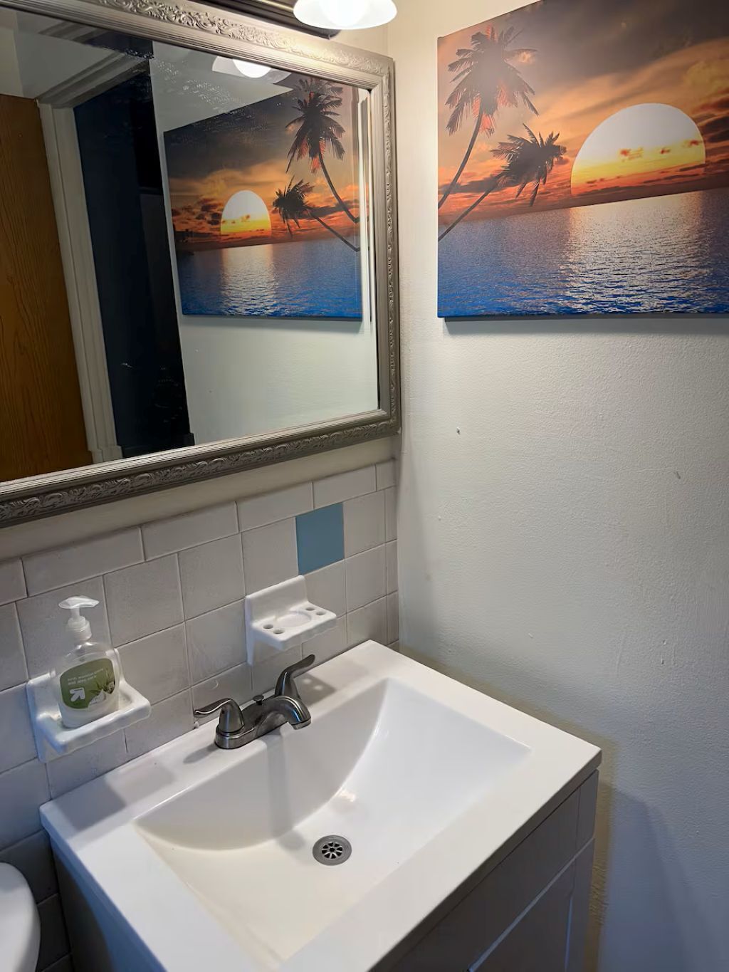 Une salle de bain avec un lavabo blanc, un miroir et deux tableaux représentant un coucher de soleil accrochés au mur.