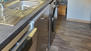 A kitchen with a brick wall, stainless steel appliances, a dishwasher, and a microwave. - Appartement de 76m2 dans maison historique (Fontaine-de-Vaucluse)
