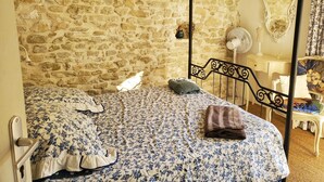 A room with a stone wall, a four-poster bed, a nightstand, a mirror, and a fan. - Appartement de 76m2 dans maison historique (Fontaine-de-Vaucluse)