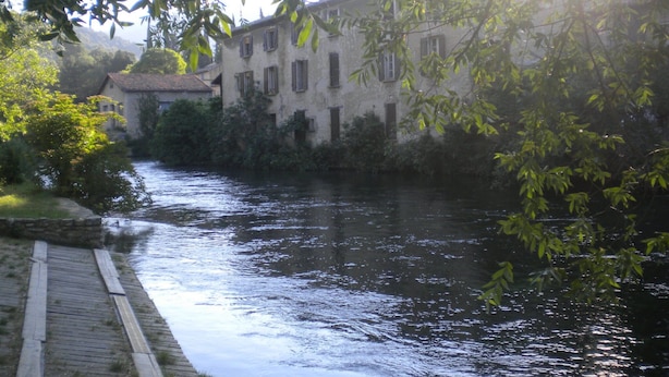 A river with a wooden walkway, buildings with multiple windows, and surrounding greenery. - Appartement de 76m2 dans maison historique (Fontaine-de-Vaucluse)