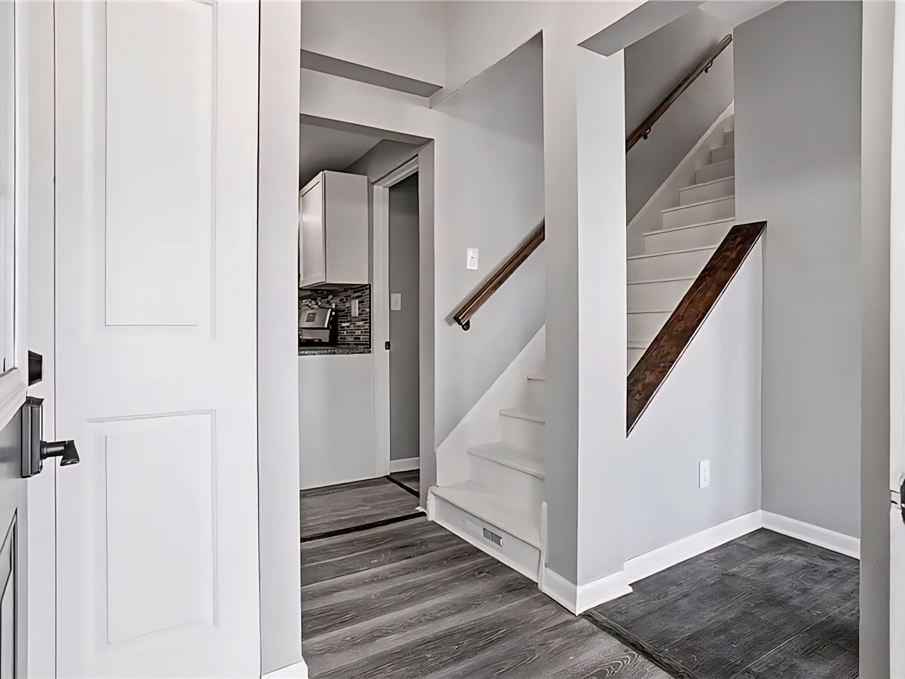 A hallway with a white door, a staircase with wooden steps, and a kitchen area in the background.
