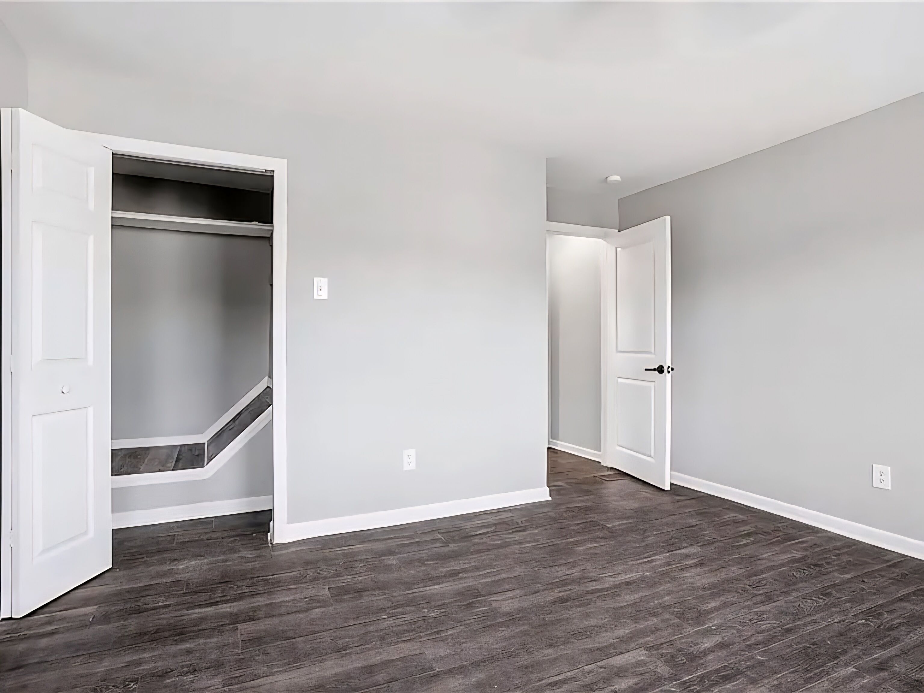 A hallway with a wooden floor, a closet with a grey door, and a white door at the end.