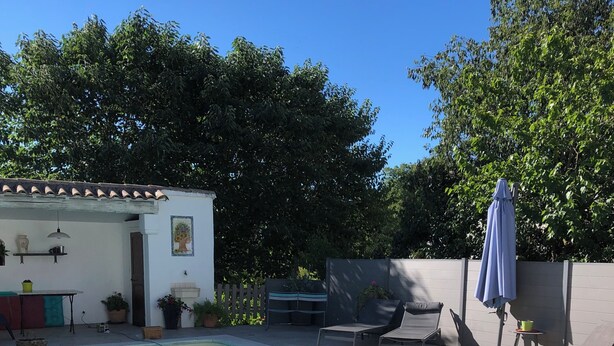 Uma piscina com deck revestido, uma pequena área de varanda com mesa e cadeiras, e um espaço de estar coberto.