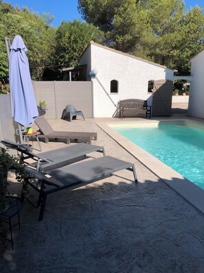 Un espace aménagé au bord de la piscine, avec des chaises longues, un parasol et un petit bâtiment doté d’une porte.