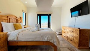 A bedroom with a bed, wooden headboard, bedside tables, a dresser, a flat-screen TV, and a view of the outdoors.