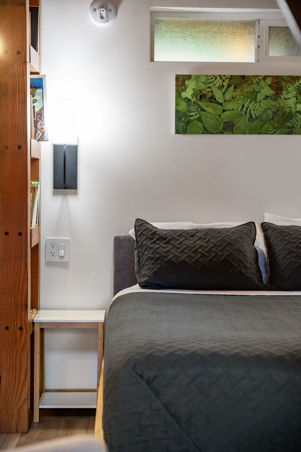 Une chambre à coucher comprenant un lit, une table de chevet, une lampe fixée au mur, une prise de courant et une œuvre d’art représentant des feuilles vertes, placée au-dessus du lit.