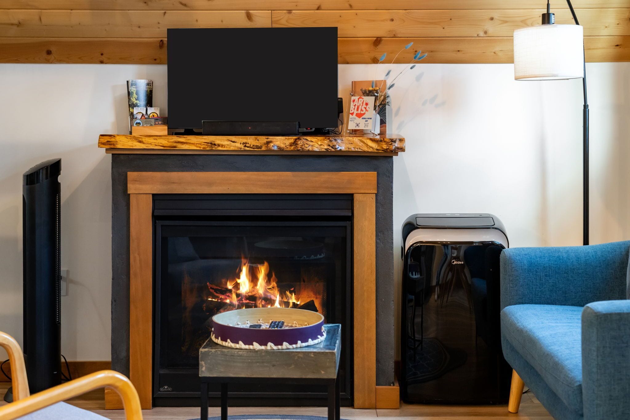 A fireplace with a burning log, a television mounted above it, and a blue chair to the right.