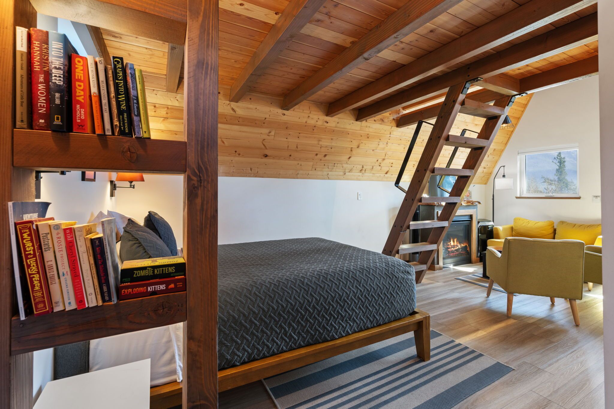 Une chambre de style « loft » avec un plafond en pente, des poutres en bois, un lit recouvert d’une housse à motifs gris, une bibliothèque remplie de livres, un fauteuil jaune et une cheminée.