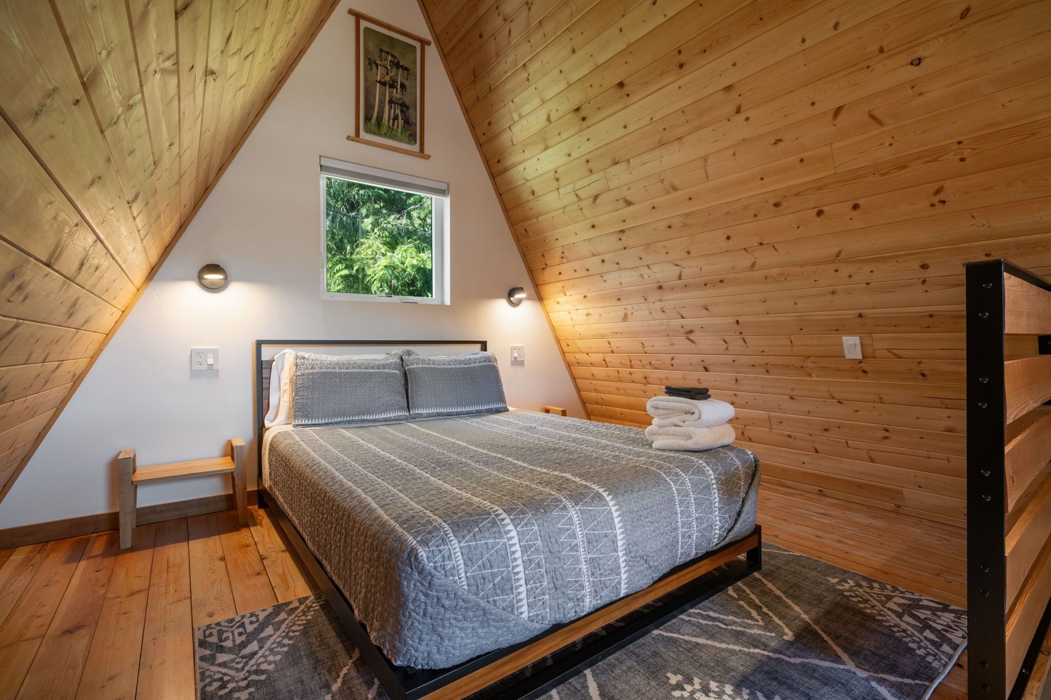 Une chambre avec un plafond en pente en bois, un lit recouvert d’une couvre-lit à motifs, une fenêtre donnant sur la verdure et un petit banc.