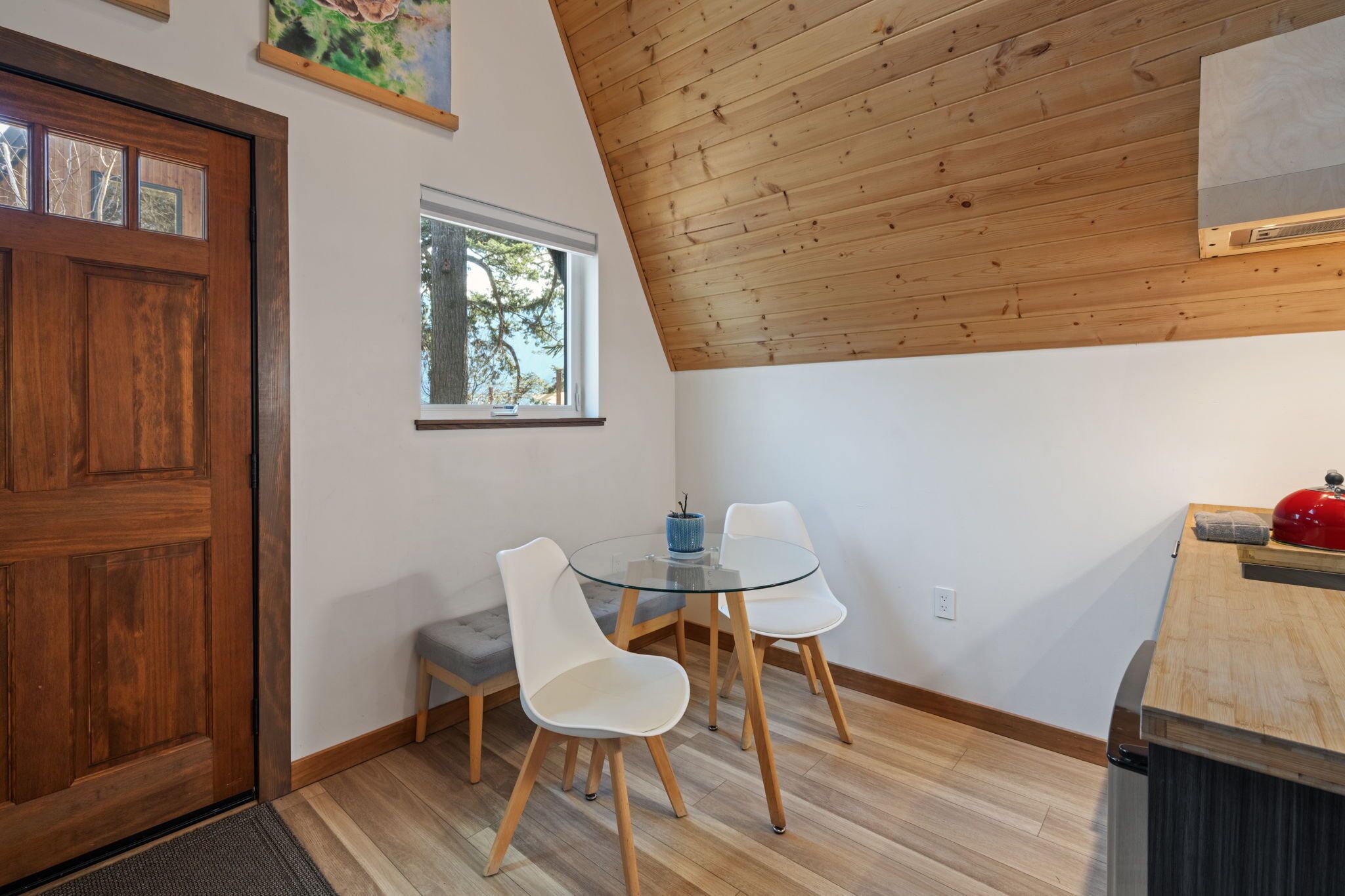Una habitación con techo de madera, una mesa de vidrio con dos sillas, una puerta de madera, una ventana y una pequeña zona de cocina con una olla roja.