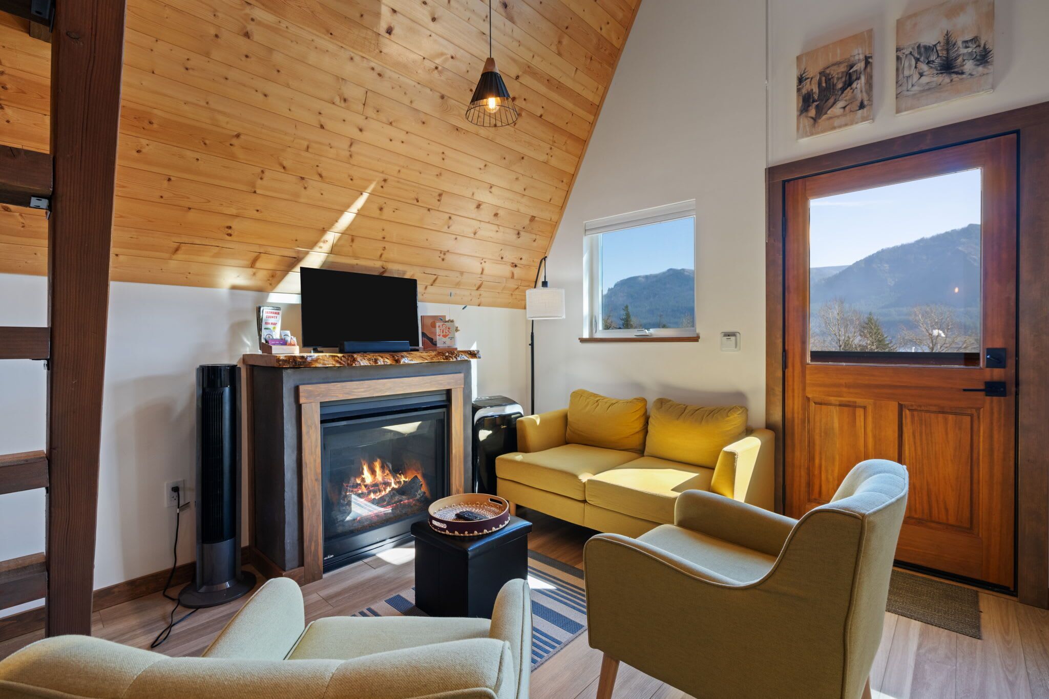 A cozy living room with a fireplace, a sofa, a TV, and a view of mountains through a large window.
