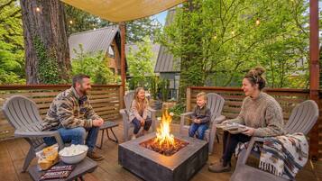 A group of people sitting around a square fire pit on an outdoor deck with chairs and a table with snacks.