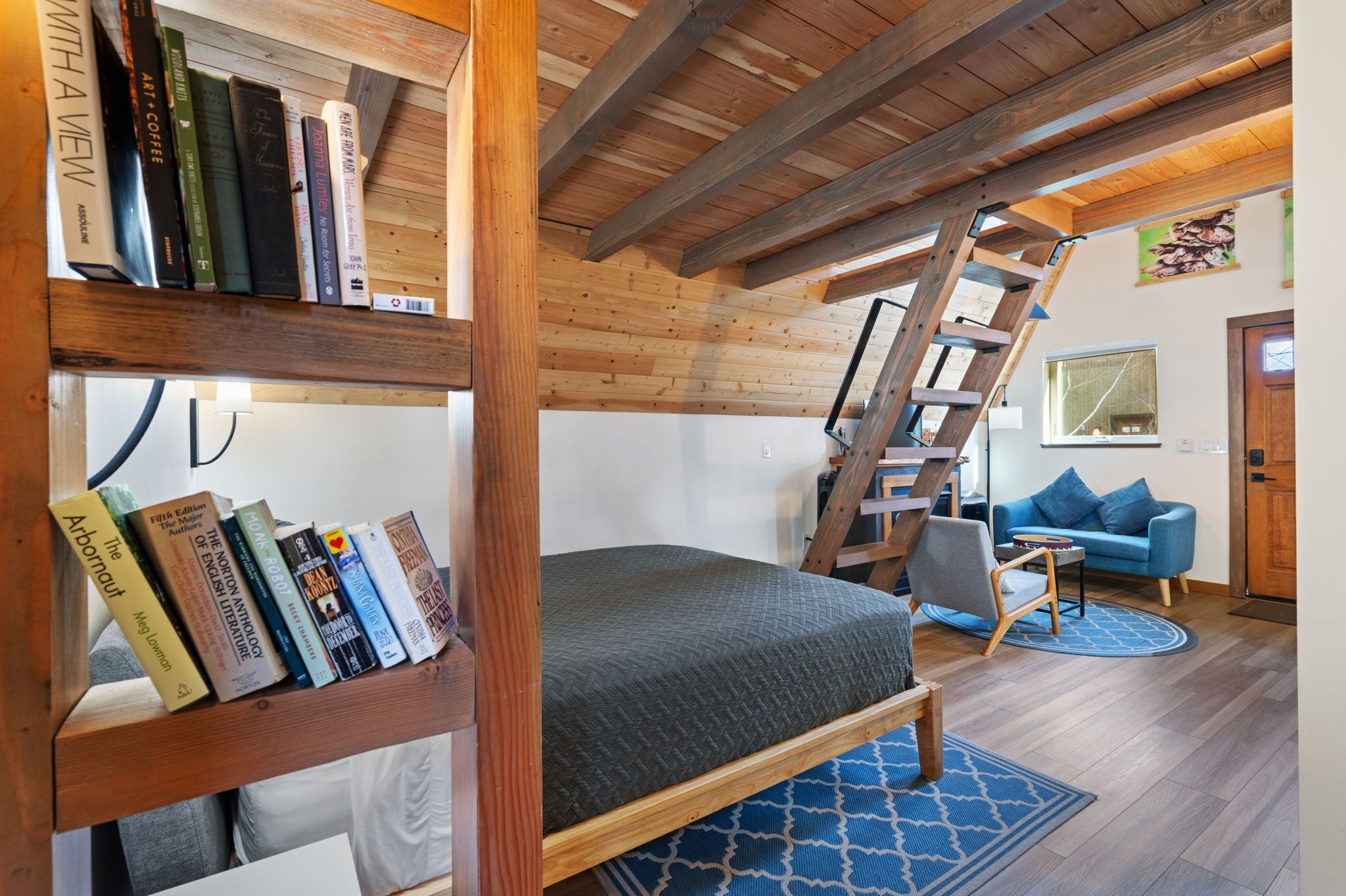 A loft-style room with a bunk bed, a bookshelf, a ladder, a blue couch, and a wooden ceiling.