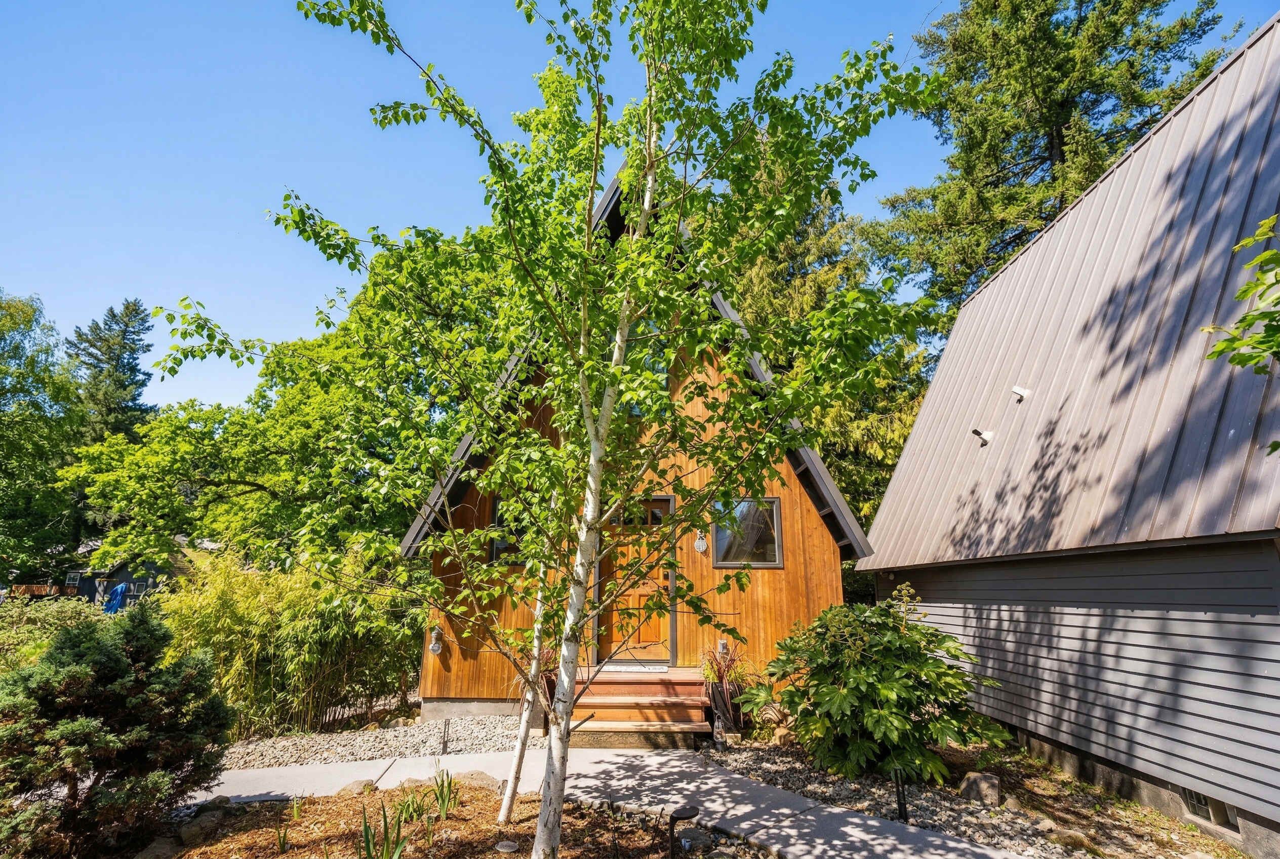 A wooden house with a sloped roof, a small porch, and a garden with various plants and trees.