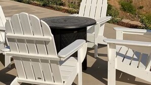A patio with white Adirondack chairs and a round table, overlooking a hilly landscape with sparse vegetation.