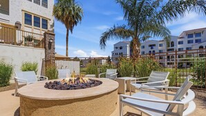A circular outdoor seating area with a fire pit, surrounded by white lounge chairs and a pool table, with a building and palm trees in the background. - The Cays at Ocotillo | Luxury Condo | Resort Pool (Chandler)