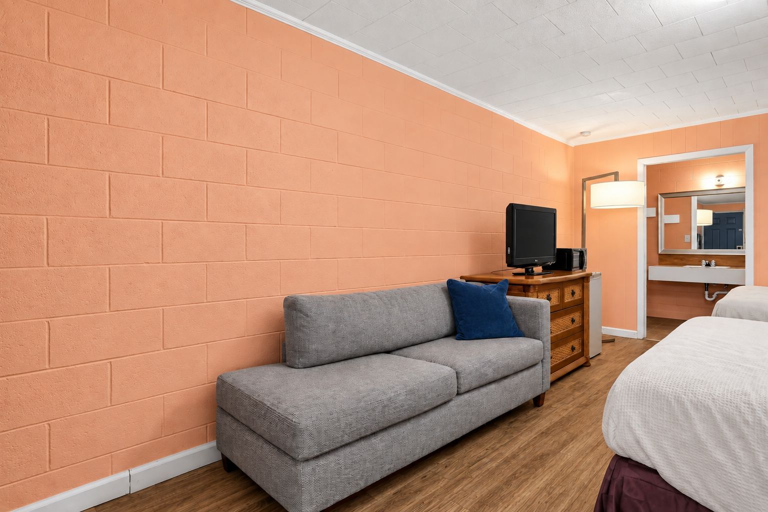 Una habitación con pared de ladrillo, un sofá gris con almohada azul, un aparador de madera, un televisor, una cama blanca y un baño con espejo y lavabo.