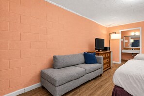A room with a brick wall, a grey sofa with a blue pillow, a wooden dresser, a television, a white bed, and a bathroom with a mirror and sink. - Budget Motel (Hamer)