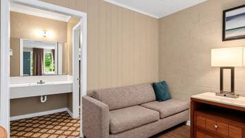 A hotel room with a sofa, a bedside table, a lamp, a mirror, and a view of the outdoors.