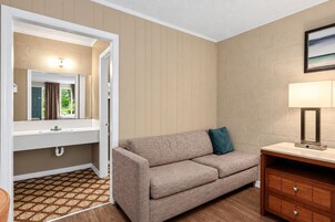 A hotel room with a sofa, a bedside table, a lamp, a mirror, and a view of the outdoors.