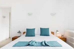 Ein ordentlich gemachtes Bett mit blauen Kissen und einer zusammengefalteten blauen Decke, eine Nachttischlampe, ein gerahmtes Bild und ein weißer Heizkörper.