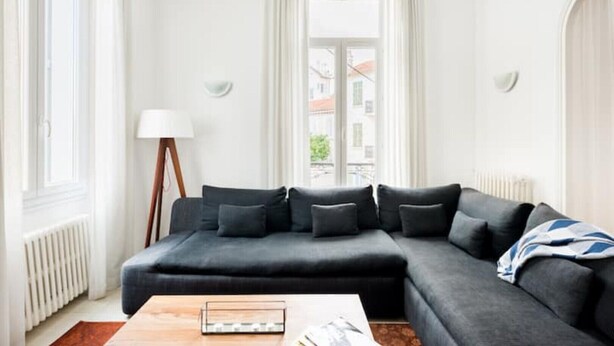 Un salon moderne avec un canapé d’angle gris foncé, une table basse en bois et une vue sur les immeubles à travers de grandes fenêtres.