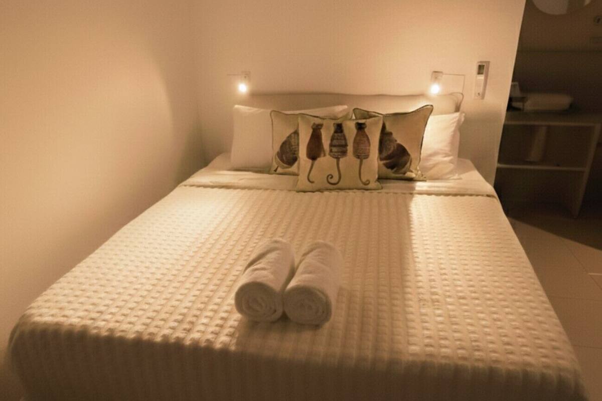 Una cama bien hecha con toallas enrolladas, un cabecero con luces empotradas y una mesita de noche con lámpara.