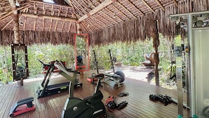 An outdoor gym area with exercise equipment, including treadmills and weights, under a thatched roof structure. - CRISTAL DA MATA (Caraíva)