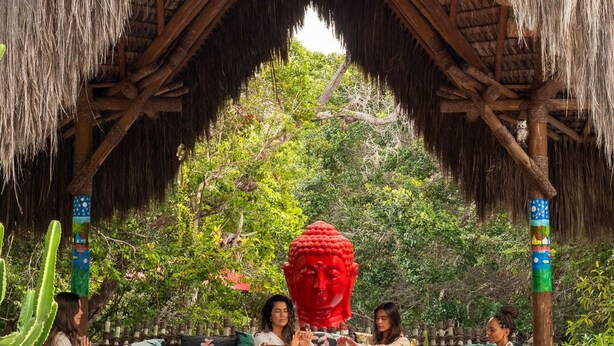 A thatched-roof structure with a Buddha statue in the center, surrounded by people engaged in a ritual. - CRISTAL DA MATA (Caraíva)