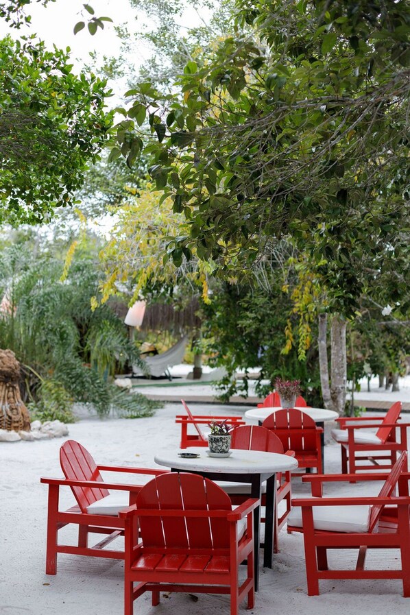 An outdoor seating area with red chairs and white tables, surrounded by greenery and trees. - CRISTAL DA MATA (Caraíva)