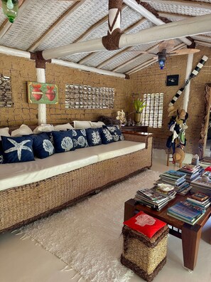 A room with a woven sofa, a small table with books, and a ceiling fan. - CRISTAL DA MATA (Caraíva)