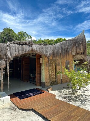 A thatched-roof structure with an open doorway, a wooden deck, and a sandy ground. - CRISTAL DA MATA (Caraíva)