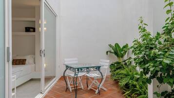 Um pequeno pátio com mesa e cadeiras, uma porta de correr de vidro que dá para um quarto e uma planta verde.