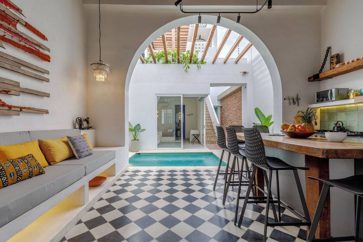 Una cocina moderna con piso a cuadros, un comedor con taburetes de bar, una banca integrada y vista a una piscina a través de un arco.