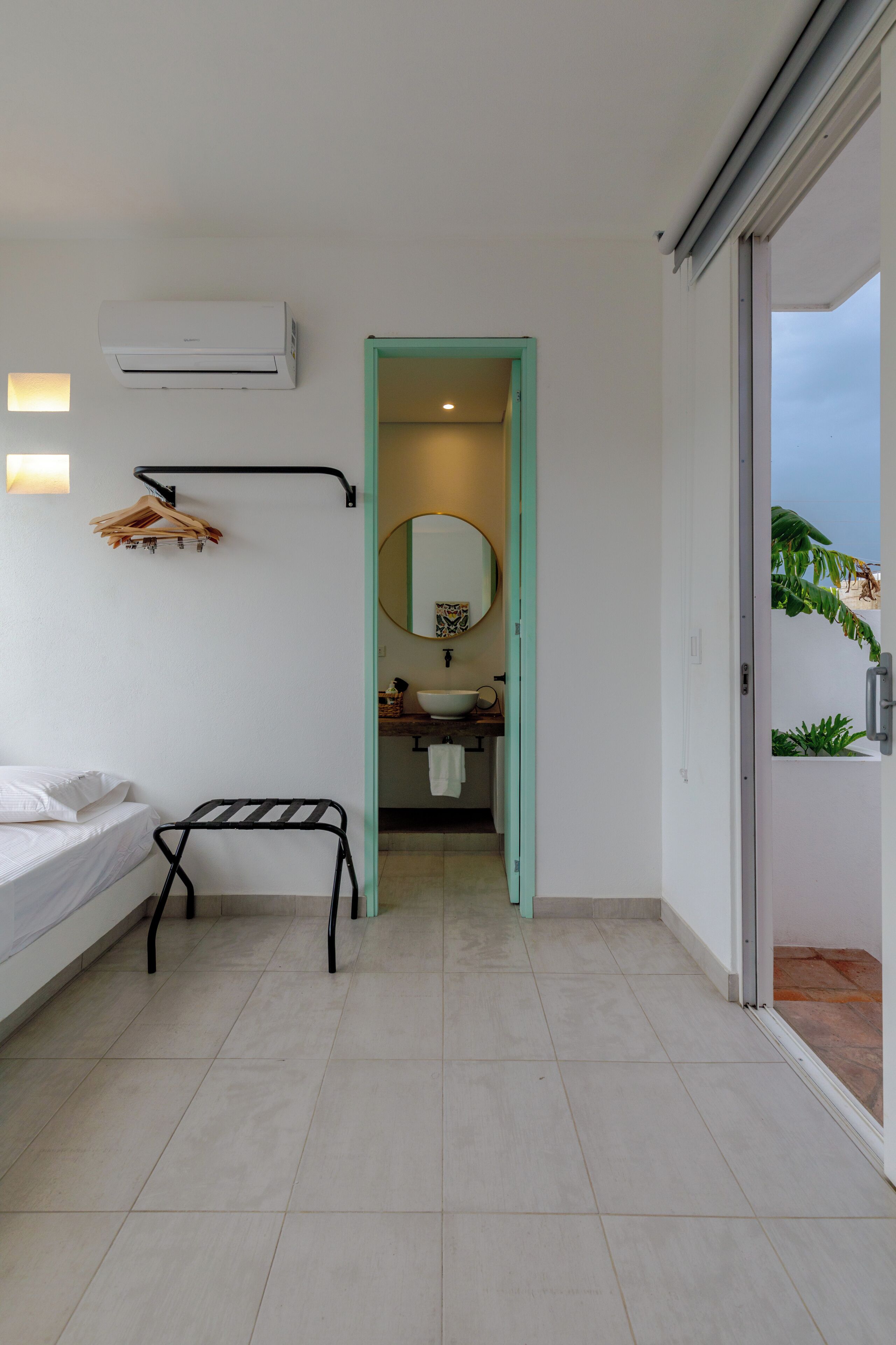 A modern hotel room with a bed, a black towel rack, an air conditioning unit, a bathroom with a sink and mirror, and a view of the outdoors.