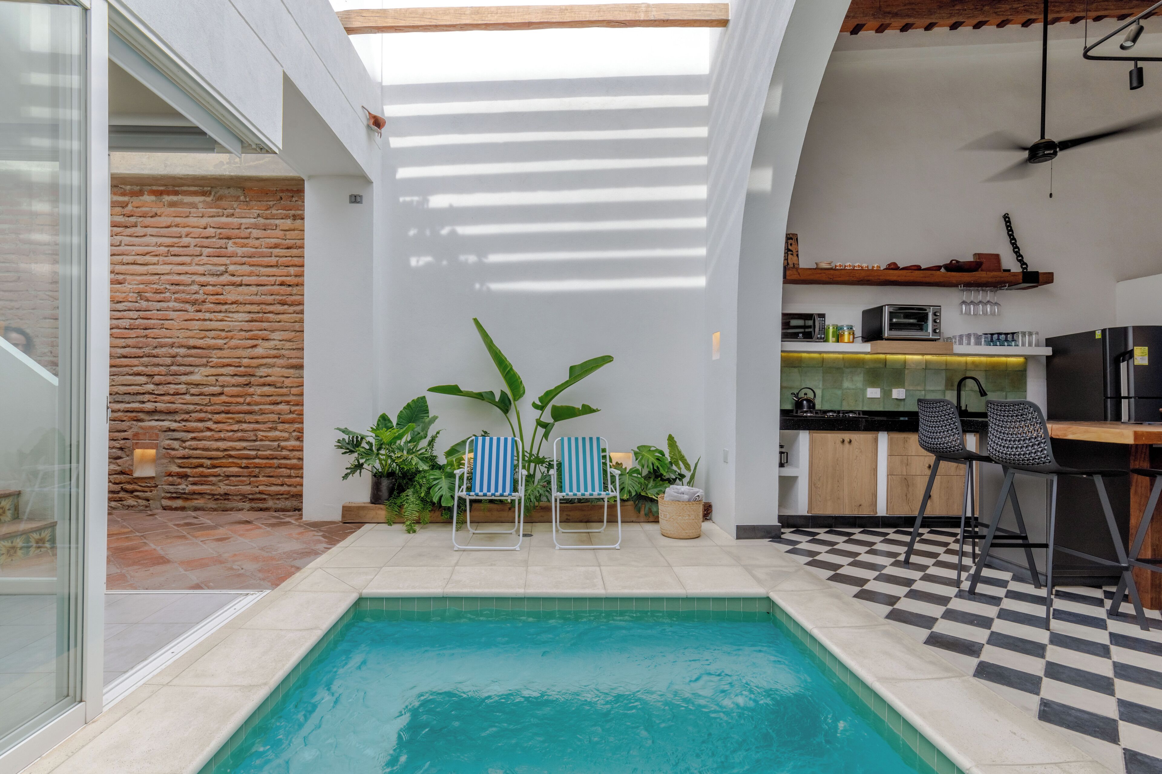 A modern indoor pool area with a checkered floor, two striped lounge chairs, a kitchen area with a microwave, and a brick wall.
