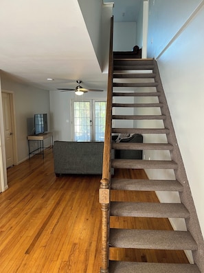 A staircase with wooden steps and a handrail, a ceiling fan, and a grey sofa in a room with wooden flooring. - Charming 3-bedroom house in lovely Jamesport- near water, Riverhead, Shinnecock. (Jamesport)