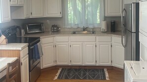 A kitchen with white cabinets, a microwave, a stove, a refrigerator, and a window with sheer curtains.