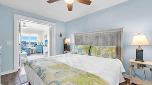 A bedroom with a bed, a wooden headboard, a nightstand with a lamp, and a view of the ocean through an open door. - Sands of Laguna B-1 (Panama City Beach)
